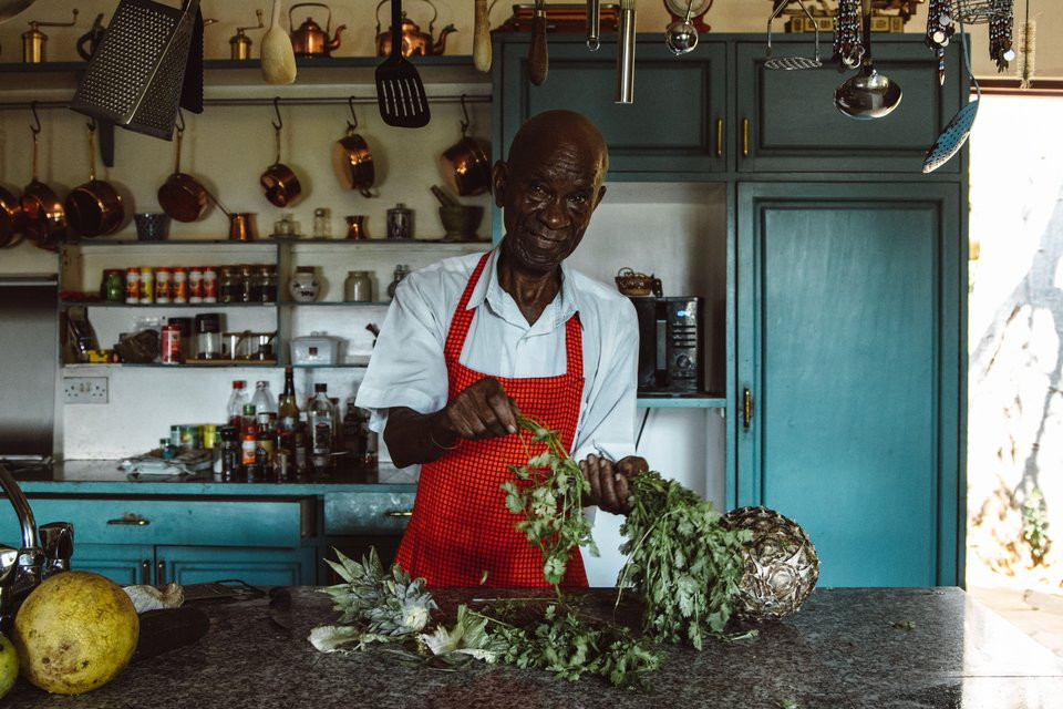 Black adult cook in an apron preparing fresh herbs at a kitchen counter.